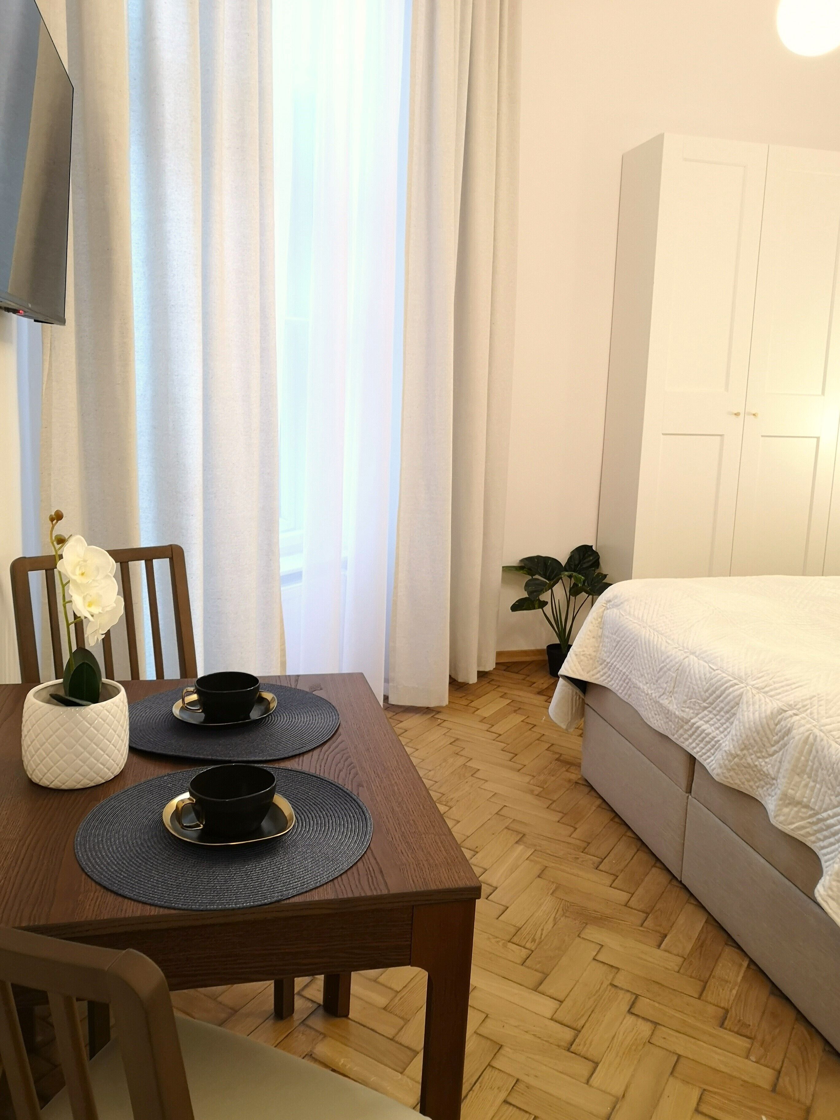 A neatly arranged room with a wooden dining table, two chairs, a white sofa, a TV mounted on the wall, and a potted plant.