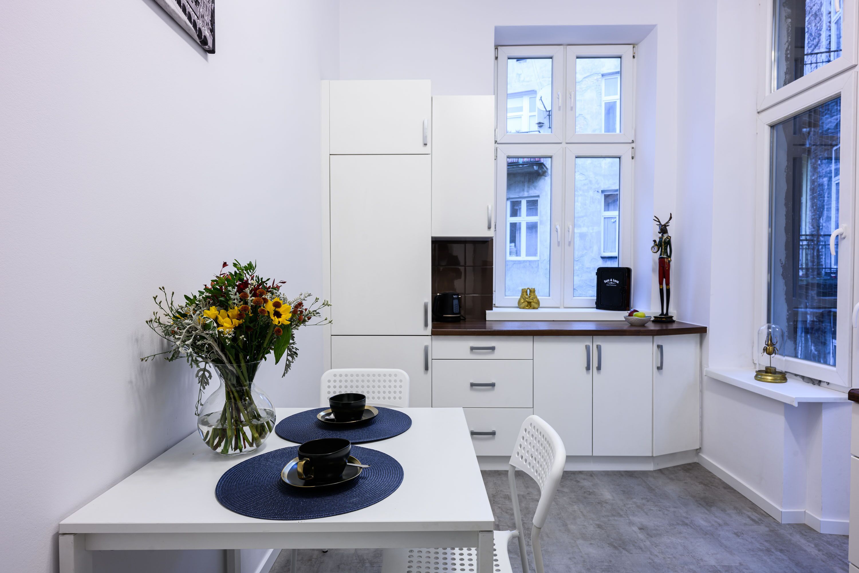 Une petite cuisine moderne avec des meubles blancs, une table blanche et une fenêtre donnant sur des immeubles.