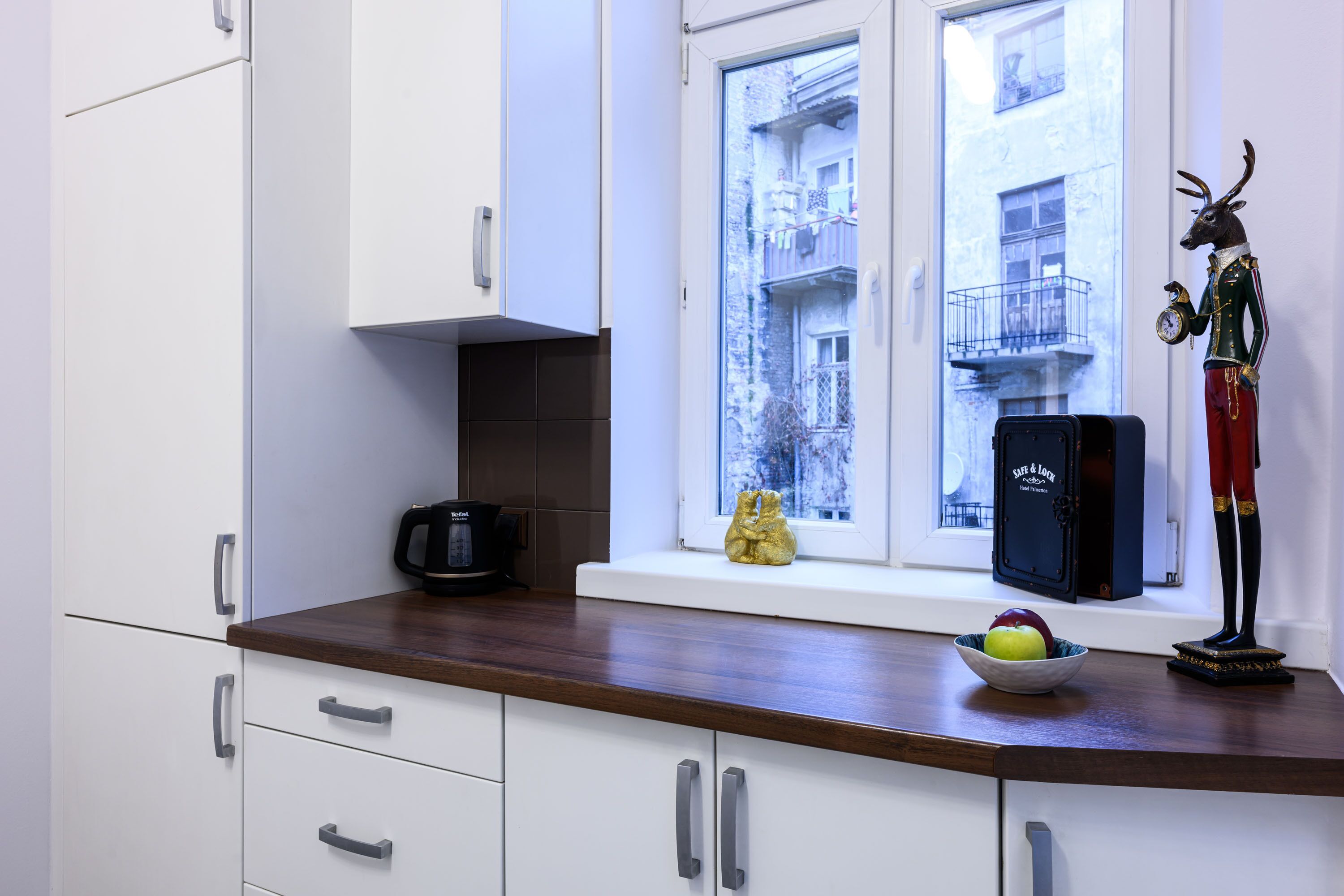 A kitchen with white cabinets, a wooden countertop, a window with a view of a building, a coffee maker, a bowl with apples, and a figurine.