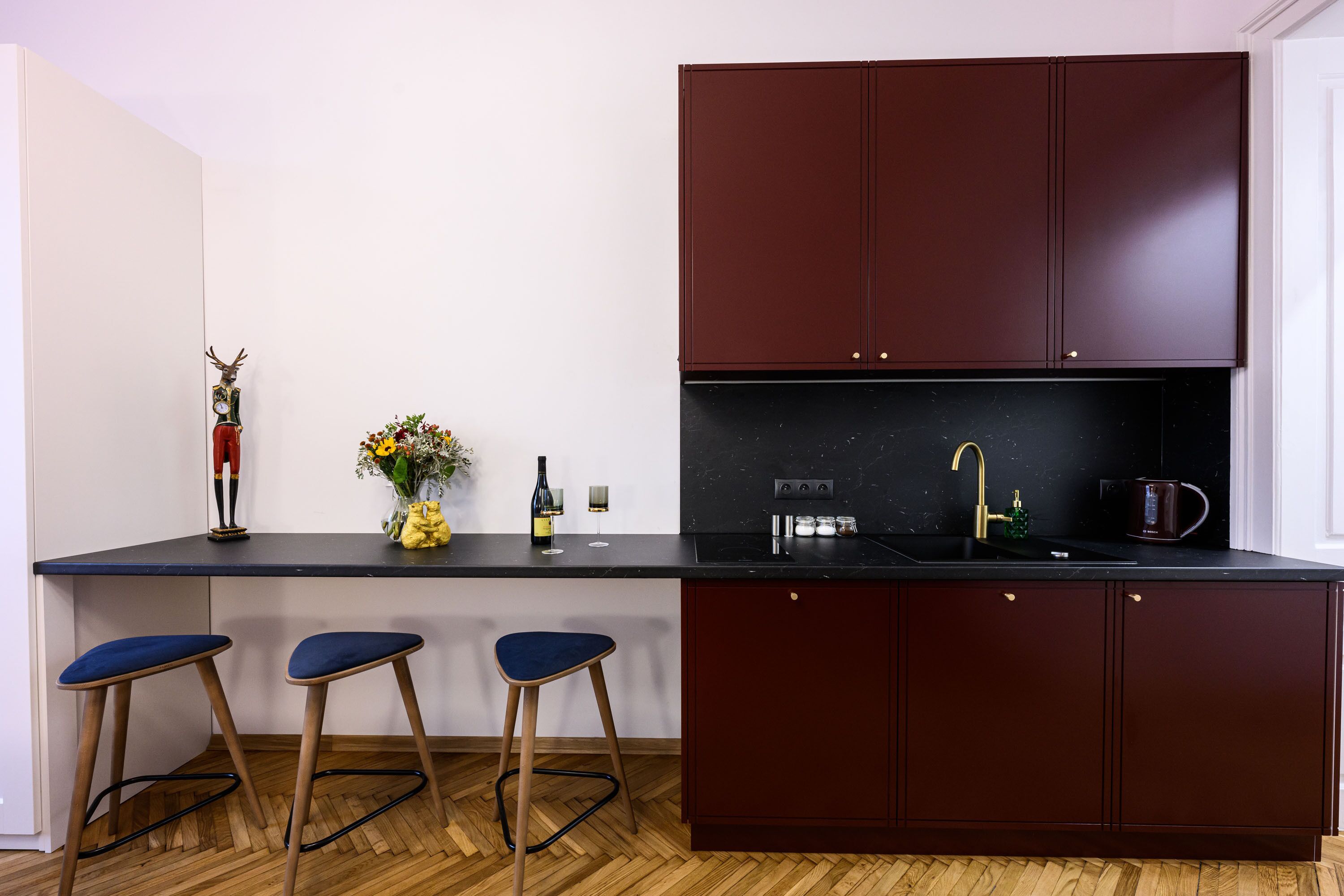 A modern kitchen with dark red cabinets, a black countertop, and three blue bar stools.