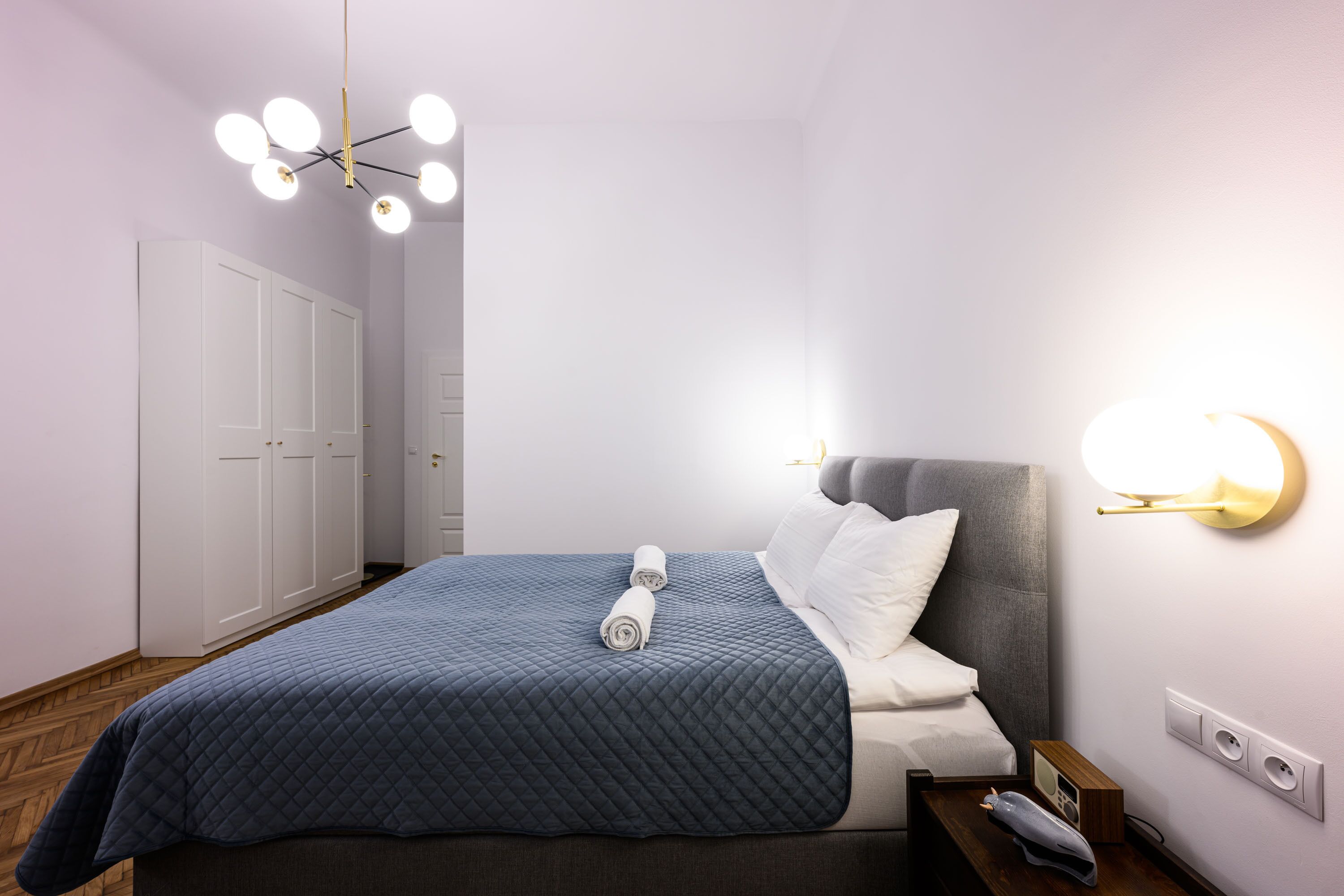 A neatly made bed with a quilted blue blanket, a grey headboard, and a bedside table with a lamp.