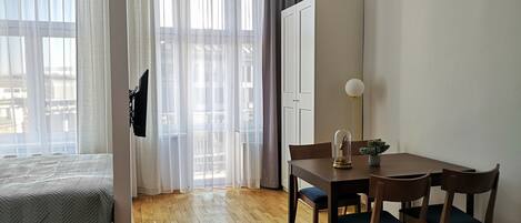A modern room with a bed, a dining table with chairs, a pendant light, and a large window with curtains.
