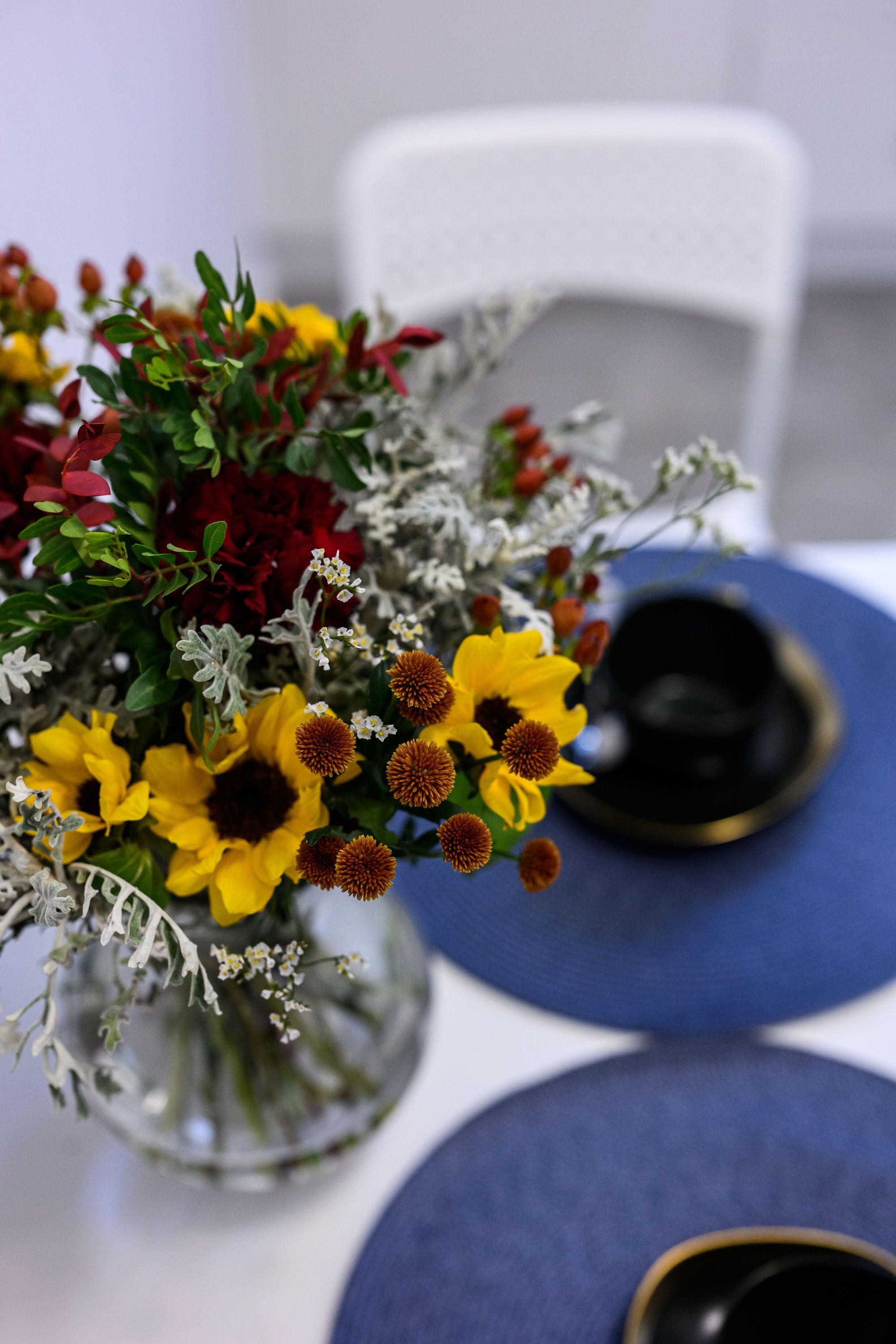 Una mesa con un florero de vidrio transparente que contiene un ramo de flores, incluyendo flores amarillas y rojas, y un objeto negro sobre la mesa.