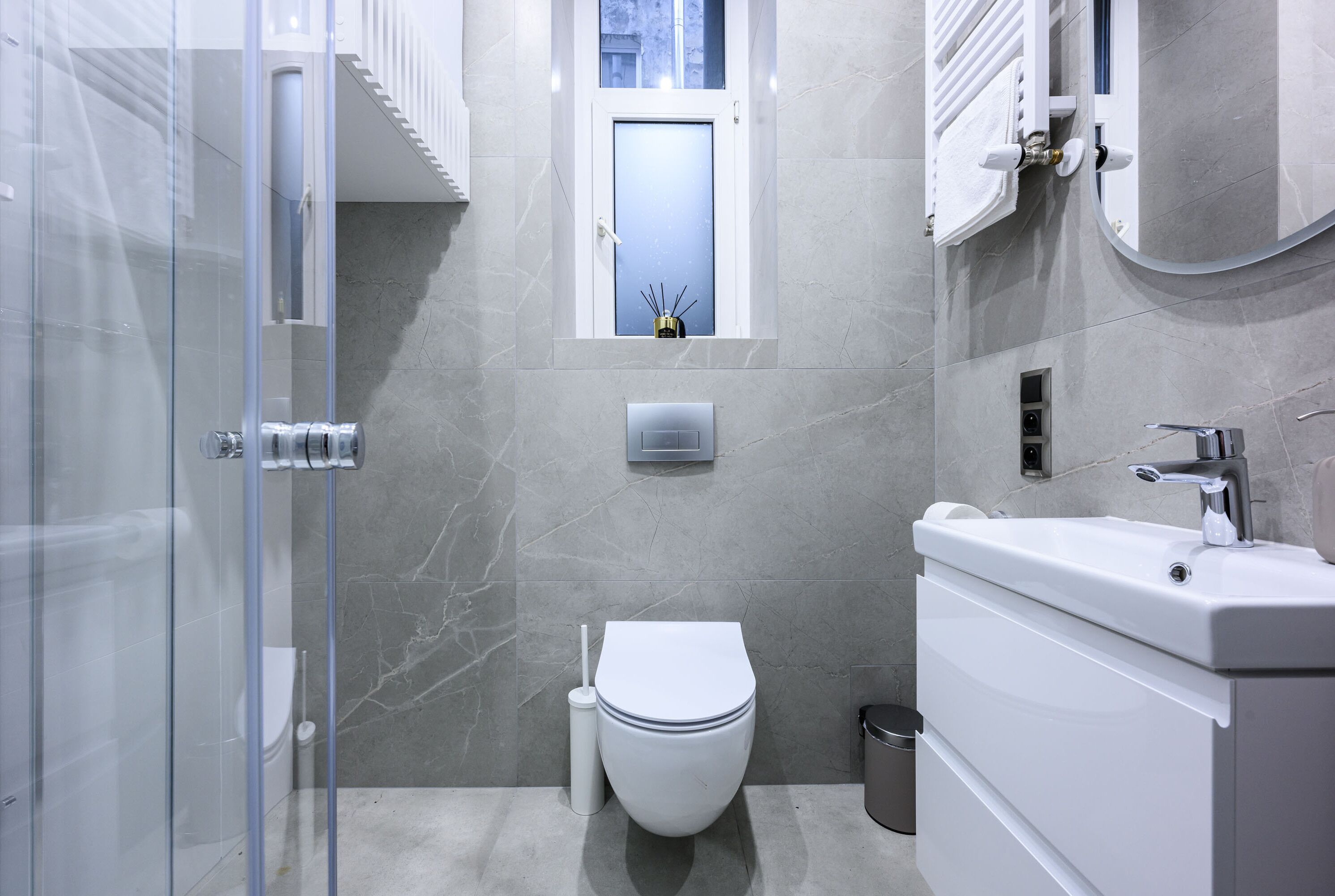 A modern bathroom with a glass shower enclosure, a toilet, a sink, and a small window above the toilet.