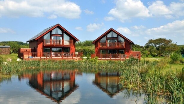 Twee rode houten huizen met balkons, weerspiegeld in stil water, omgeven door groen.