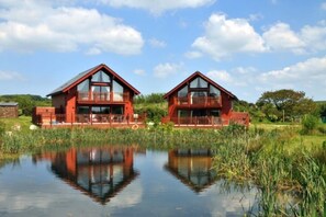 Two red wooden houses with balconies, reflecting in a calm body of water, surrounded by greenery. - Puffin lodge 4 bedroom with Hot tub,-Indoor Pool & Gym (Padstow)