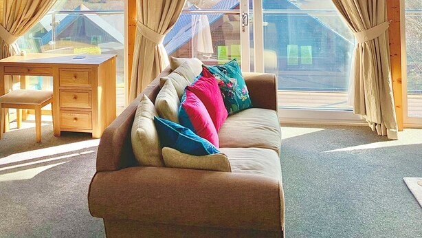 Ein Zimmer mit Holzboden, einem Sofa mit bunten Kissen, einem Holztisch und großen Fenstern mit Vorhängen.
