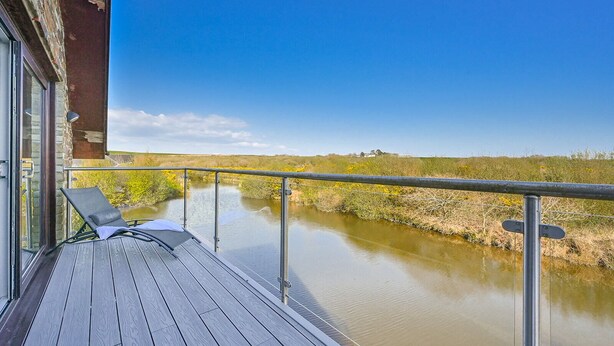 A wooden deck with a glass railing overlooking a calm river, a lounge chair, and a building with a stone facade.