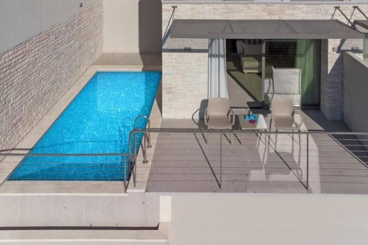 Piscina no terraço com um recurso de água azul cristalina, cercada por um muro de pedra e área coberta com assentos, incluindo duas cadeiras e uma mesa.