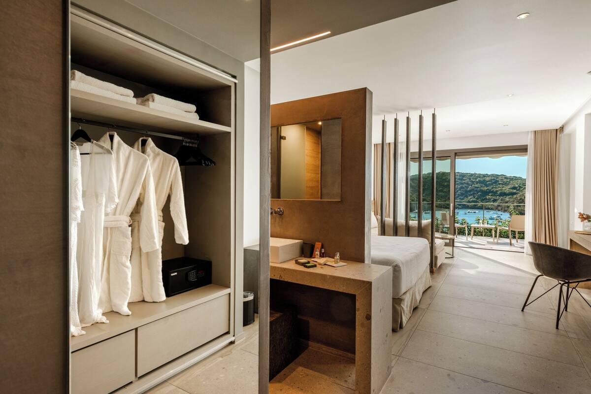 Quarto de hotel moderno com um guarda-roupa grande, casa de banho com lavatório e espelho, cama e varanda com vista para o mar.