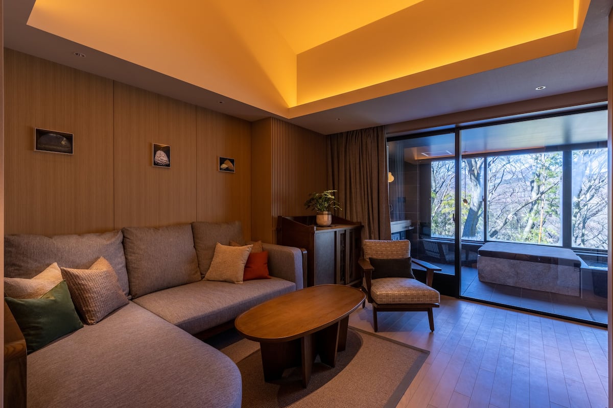 A modern living room with a sofa, armchair, and coffee table. There is a large window with a view of trees.