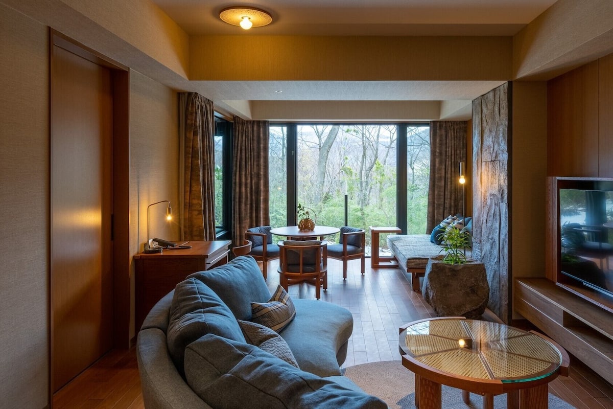 A modern living room with a sofa, dining table, chairs, a TV, and large windows.