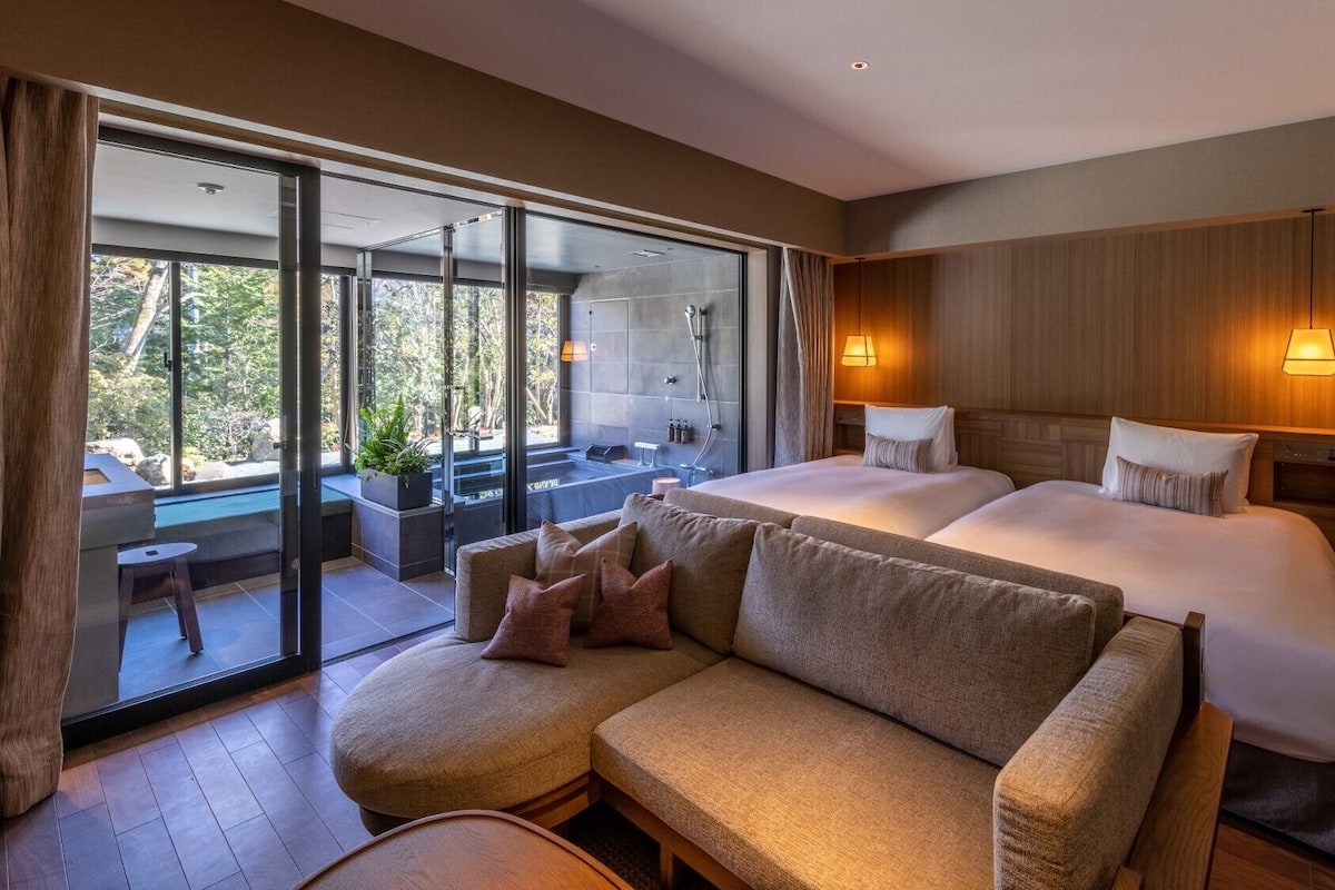 A modern hotel room with a large bed, a sofa, a glass door leading to a balcony, and a view of the outdoors.
