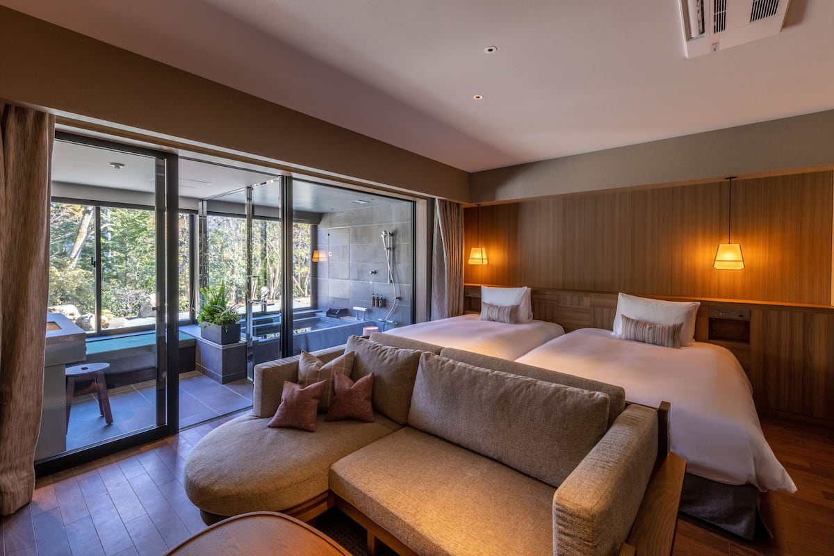 A modern hotel room with a large bed, a sofa, a glass door leading to a balcony, and a shower area.