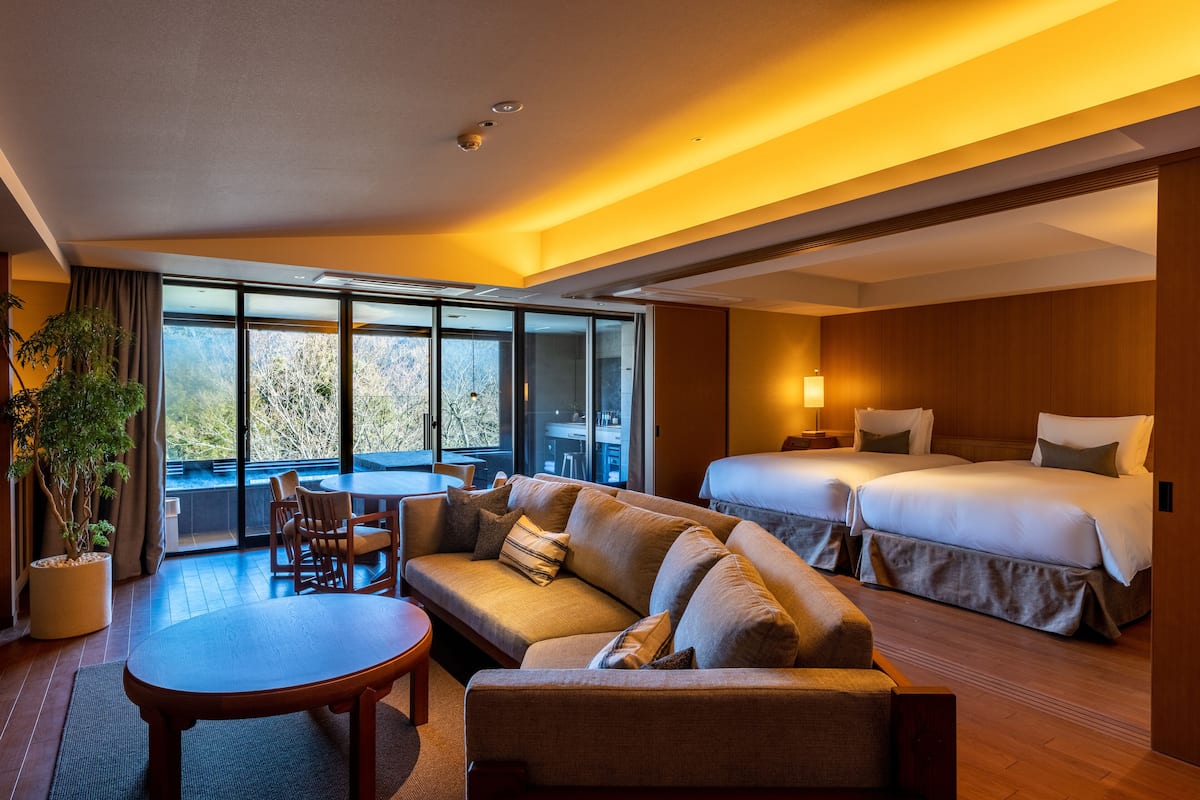 A hotel room with a sofa, a dining table, a bed, and a large window offering a view of trees.