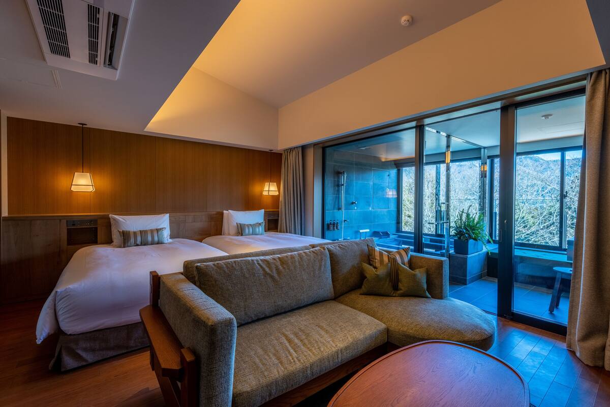 A modern hotel room with a large bed, a sofa, a round table, and a balcony with a view of the mountains.