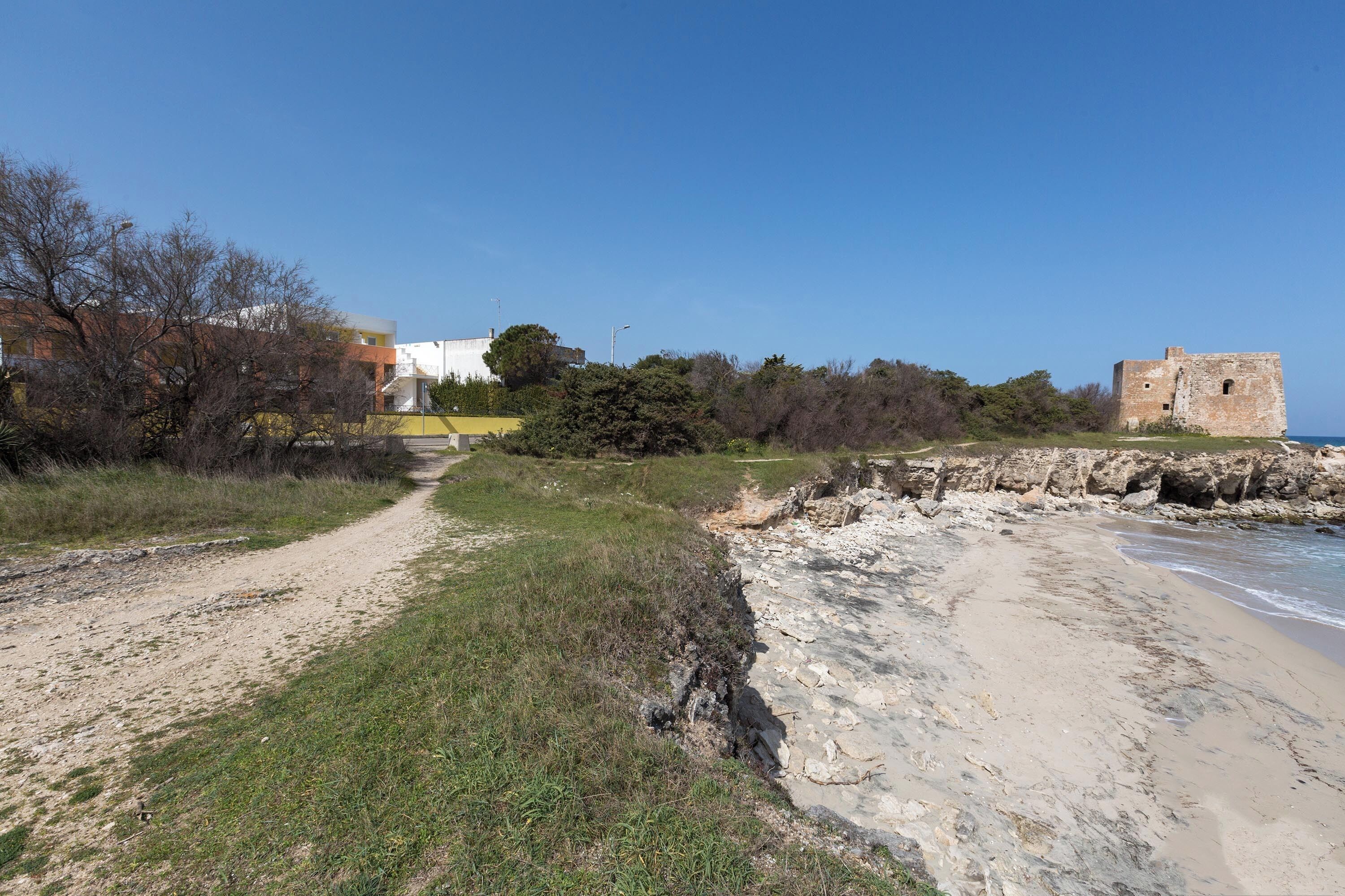 A coastal path leading to a beach with a rocky shoreline, a small tower, and buildings in the background.