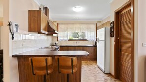 A kitchen with wooden cabinets, a built-in stove, a refrigerator, and a window with curtains. - Family Escape | Lakes Entrance Retreat (Lakes Entrance)