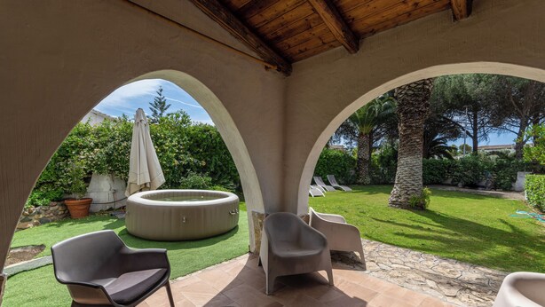A covered patio with a stone floor, two chairs, a small table, and a hot tub.