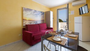 A compact living space with a red sofa, a dining table with chairs, a TV mounted on the wall, and a refrigerator.