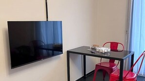 Een kleine kamer met een flatscreen televisie aan de muur, een tafeltje met enkele spullen erop, en twee rode stoelen.