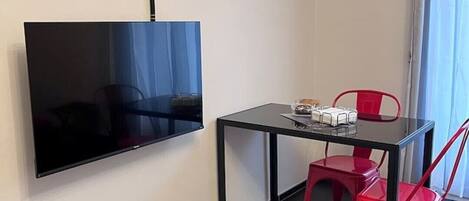 Ein kleines Zimmer mit einem an der Wand befestigten Flachbildfernseher, einem kleinen Tisch mit einigen Gegenständen und zwei roten Stühlen.