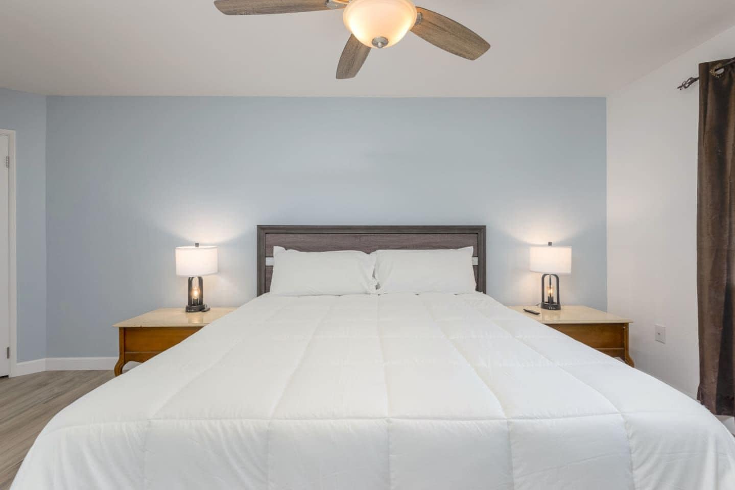 A bedroom with a single bed, two bedside tables with lamps, a ceiling fan, and a plain wall.