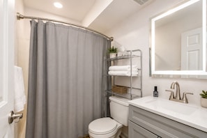 A bathroom with a shower curtain, a toilet, a sink with a mirror, and a shelf with towels.
