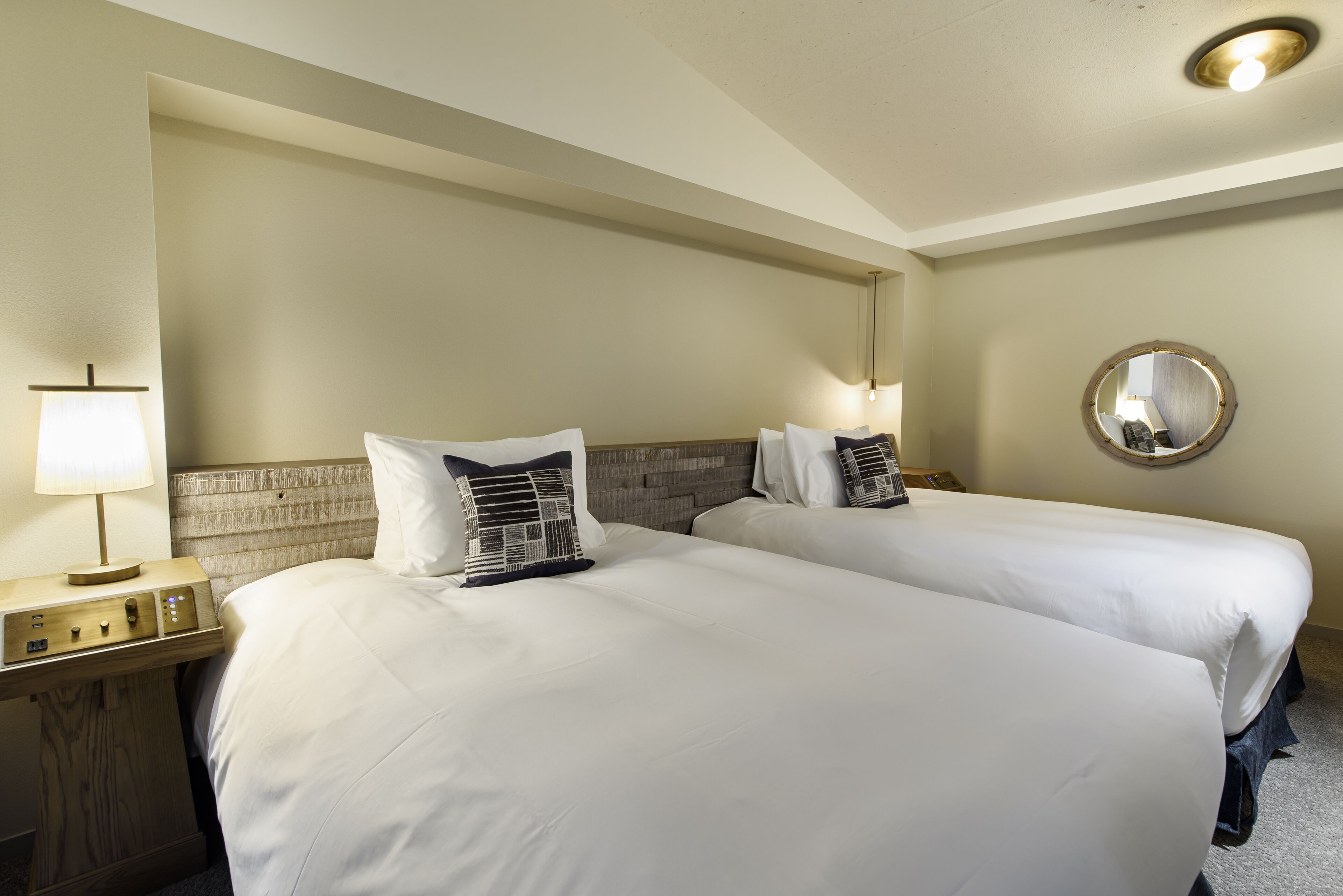 A hotel room with two beds, a nightstand, a lamp, and a mirror.