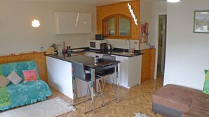 En kompakt opholdsstue med tekøkken, sofa, spisebord og lille køkkenø.