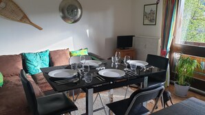 Et spiseområde med et bord dækket til fire personer, en sofa, et fjernsyn og et vindue med gardiner.
