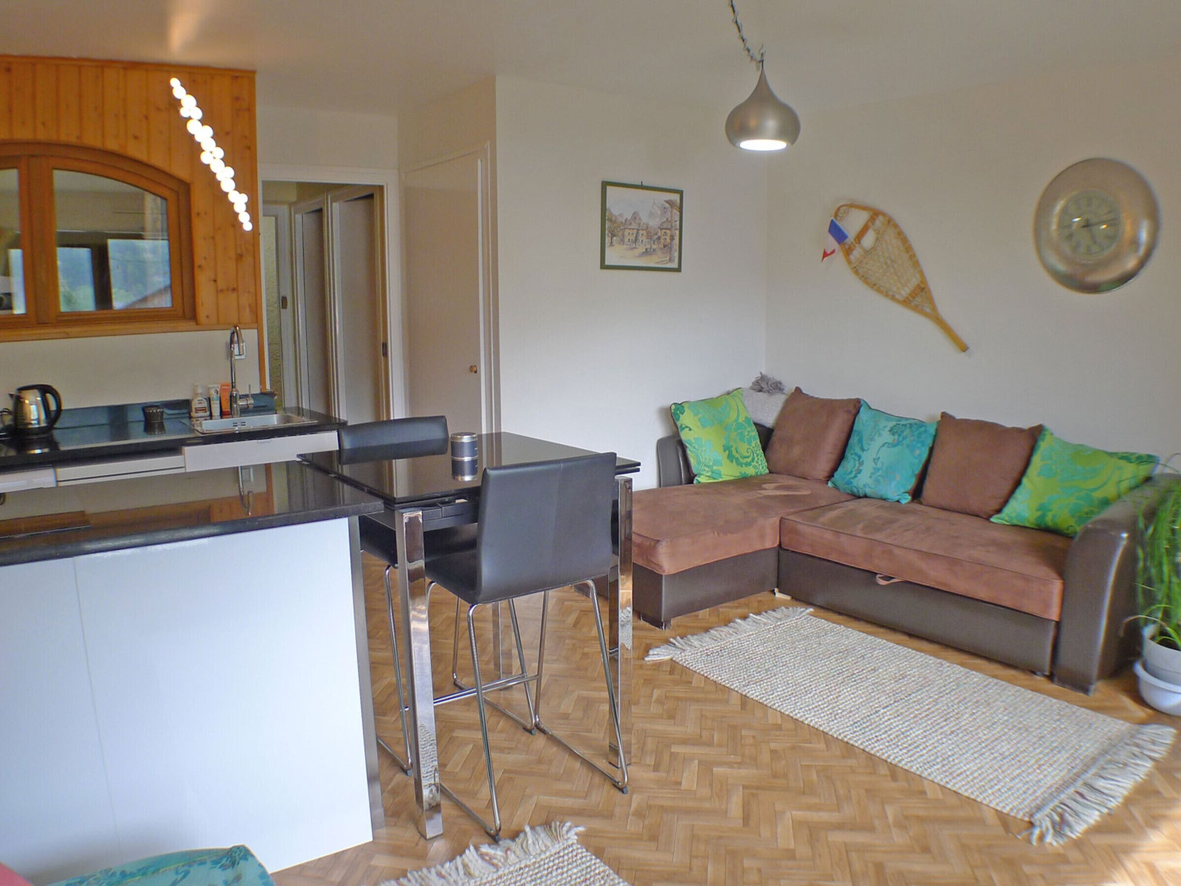 A modern kitchen with a dining area, a brown leather sofa with colorful cushions, and a wooden herringbone floor.