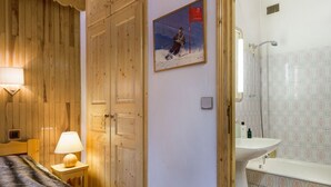 Une pièce aux murs lambrés, un lit recouvert d’un plaid en fourrure, un tableau représentant un skieur, une salle de bain avec un lavabo et un miroir, et une douche.