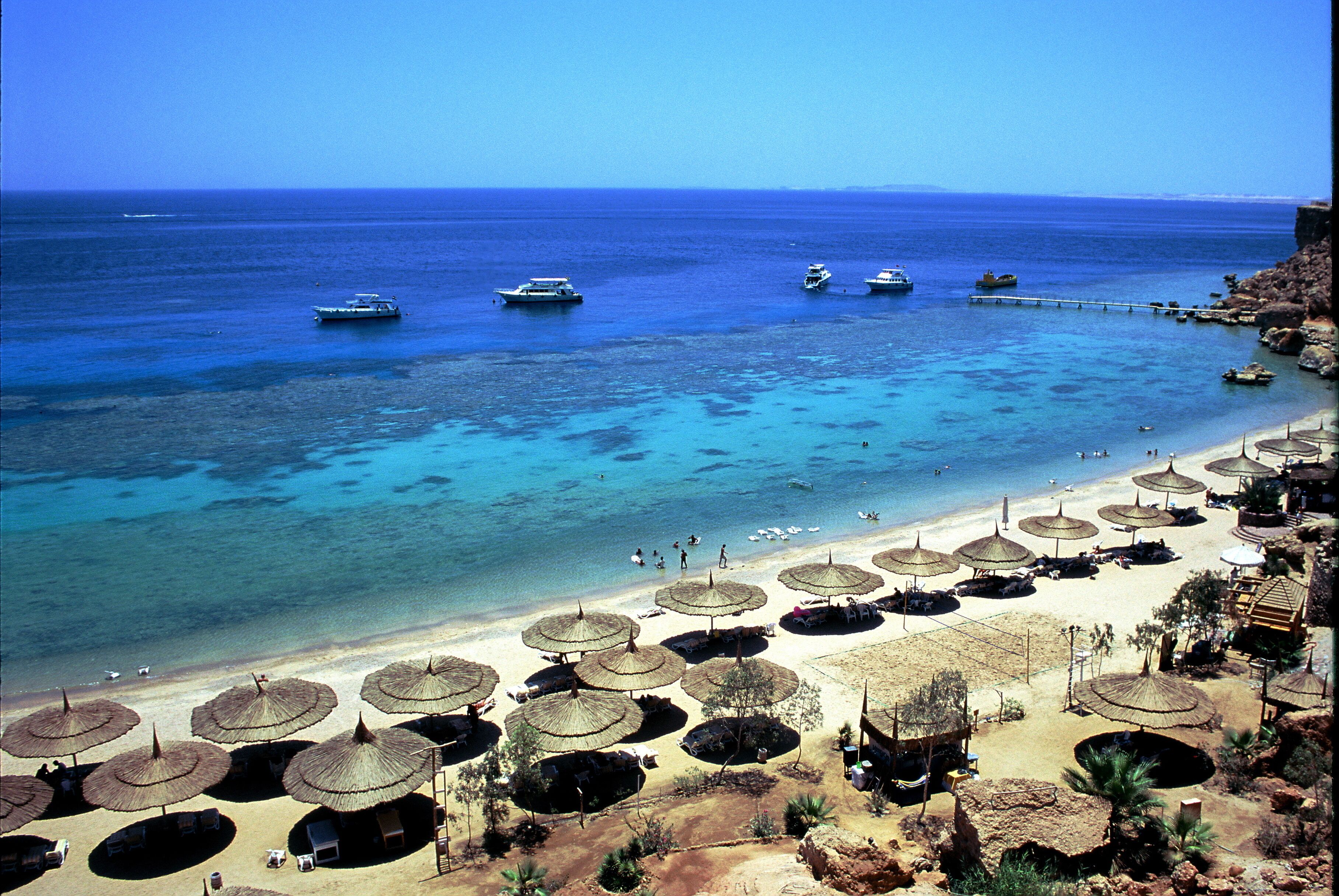 Une plage avec une eau d’un bleu limpide, plusieurs bateaux et de nombreux parasols en chaume offrant de l’ombre.