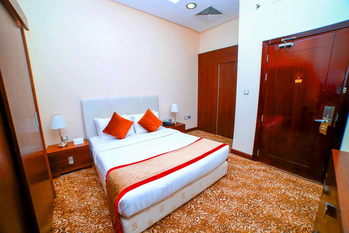 Una habitación de hotel con una cama grande, un armario de madera, una mesita de noche con lámpara y una alfombra estampada.