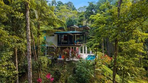 A two-story house with a balcony, surrounded by dense forest.