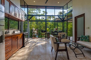 A modern kitchen with a large window, a dining area with a table and chairs, and a view of lush greenery.