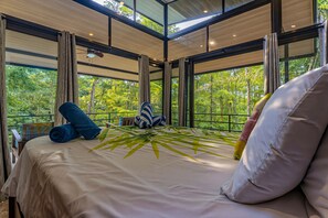 A bedroom with a large bed, a view of a forest through large windows, and a ceiling fan.