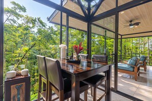 A modern outdoor dining area with a glass enclosure, wooden furniture, and a view of lush greenery.