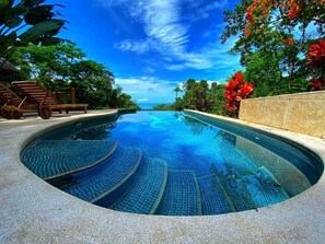 A swimming pool with a curved edge, surrounded by lush greenery and a clear blue sky. - Ocean views-2 homes-2 swimming pools-5 min to Uvita-Sleeps 11 (Uvita)