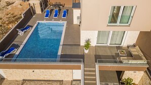 Un área de piscina en la azotea con sillones reclinables azules, una piscina azul cristalina y un edificio moderno con techo plano y grandes ventanales.