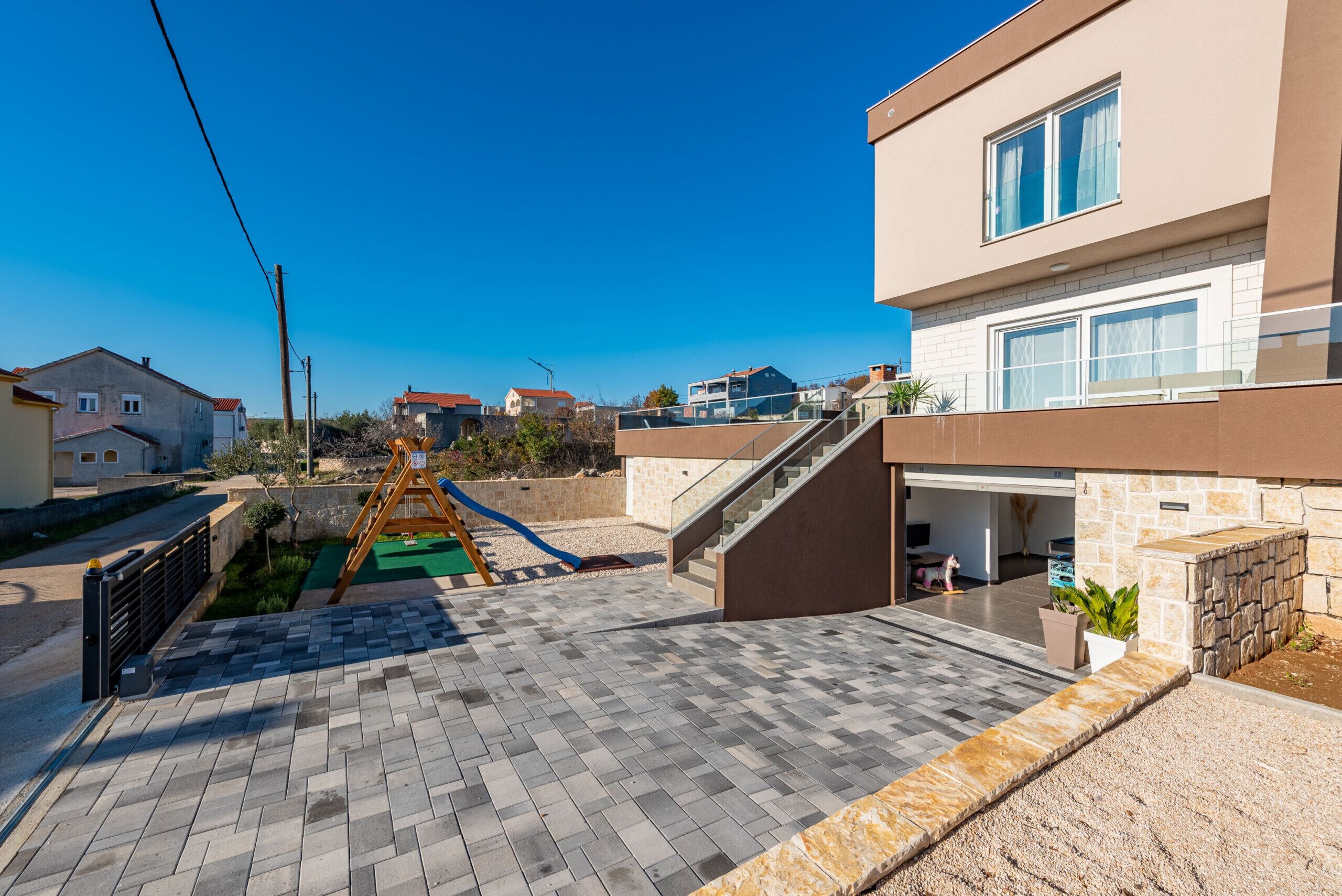 A residential area with a paved patio, a wooden swing set, and a modern house with a garage.