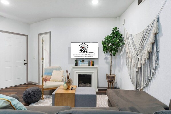 Un salon avec une cheminée, un téléviseur fixé au mur, une décoration murale en macramé et une plante en pot.