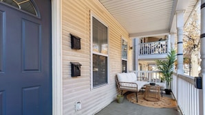 A covered porch with a white railing, a wicker chair, a small table, and a potted plant.