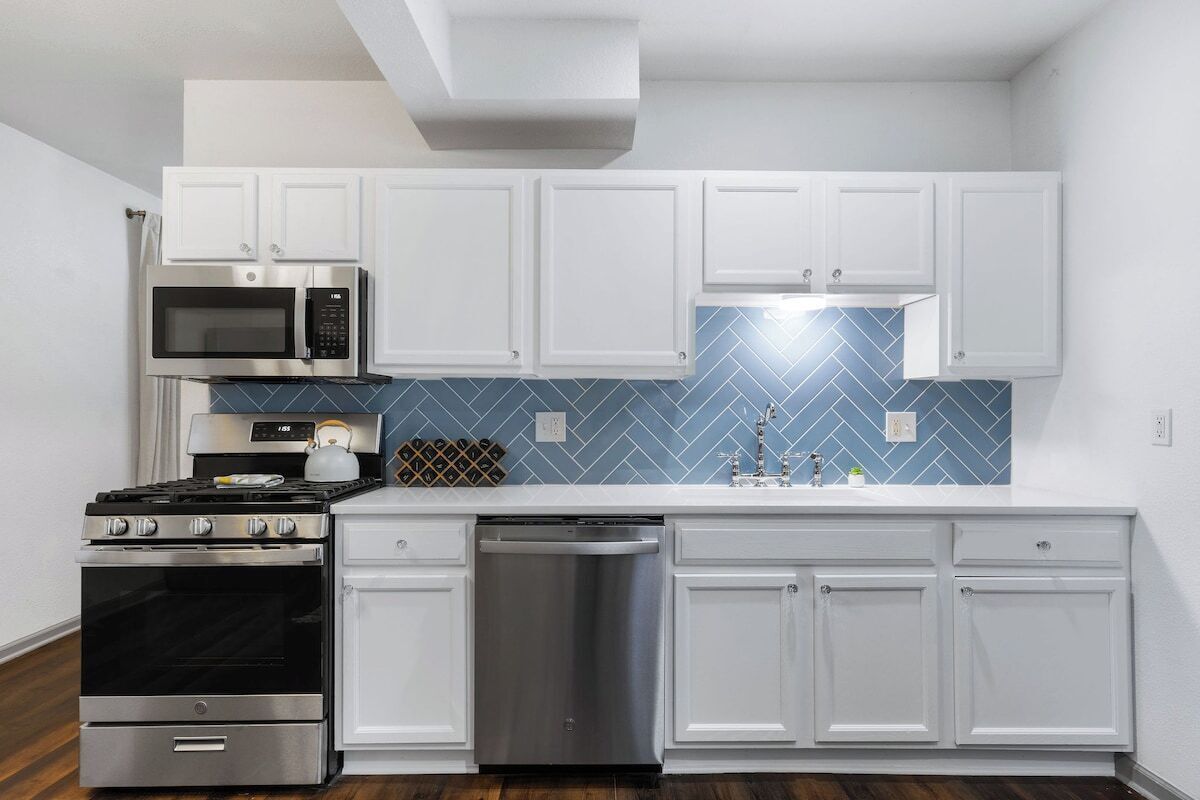 A modern kitchen with white cabinetry, stainless steel appliances, and a blue herringbone backsplash.