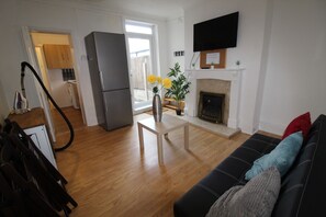 A living room with a black sofa, wooden flooring, a flat-screen TV mounted above a fireplace, and a kitchen visible through an open door.