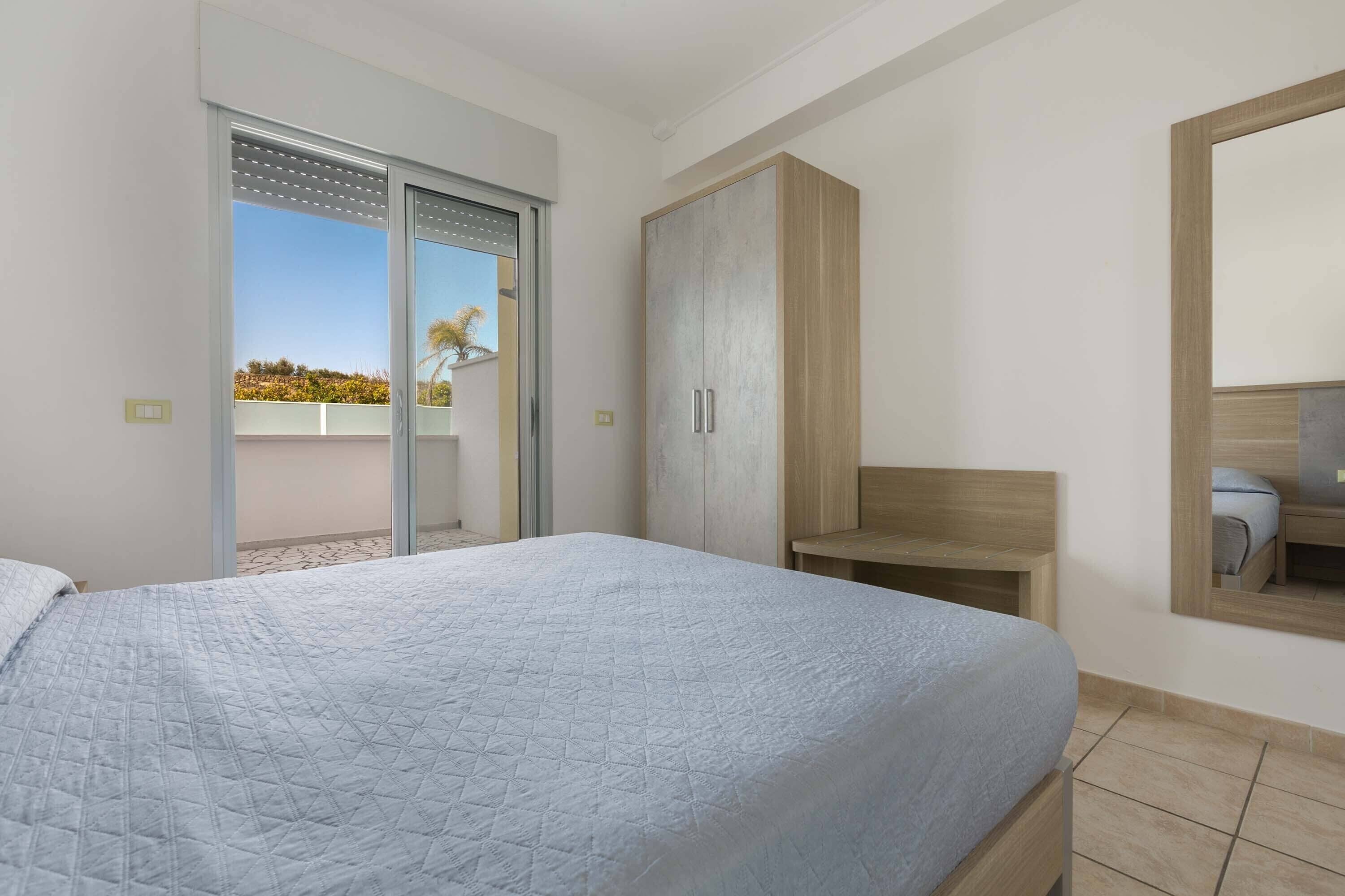 A bedroom with a bed, a wardrobe, a bathroom mirror, and a sliding door to a balcony.