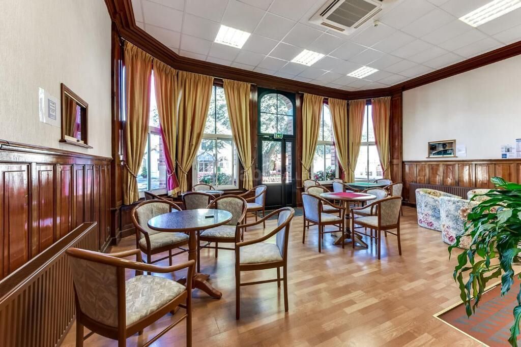 Eine Hotellobby mit Holzwandverkleidung, mehreren Tischen und Stühlen, großen Fenstern mit Vorhängen und einer Pflanze in der Ecke.