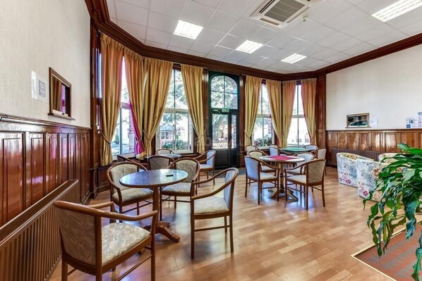 Un vestíbulo de hotel con paneles de madera, varias mesas y sillas, ventanas grandes con cortinas y una planta en la esquina.