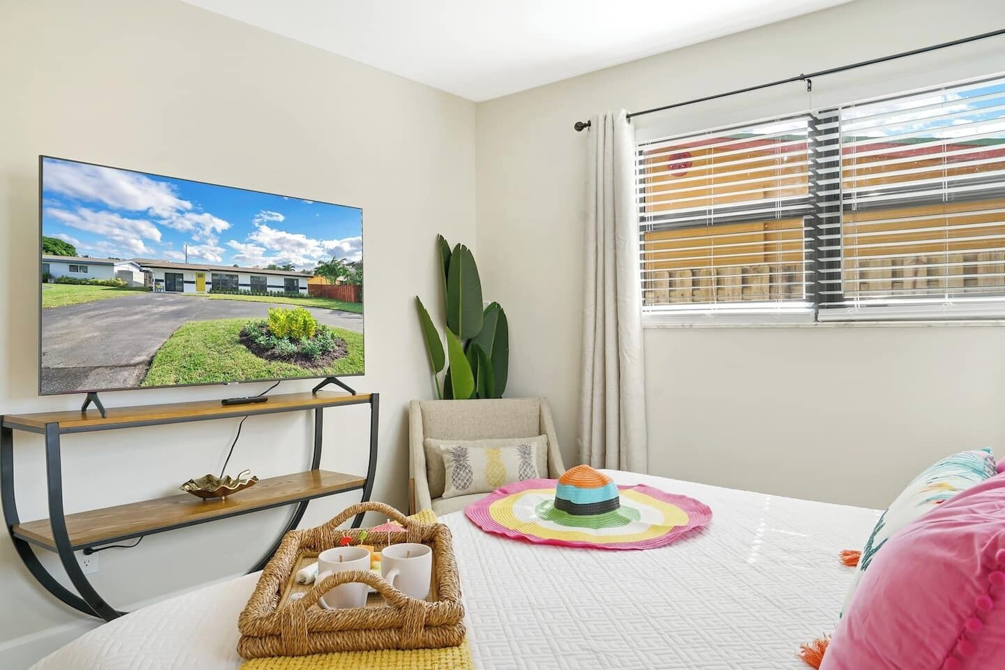 Een slaapkamer met een bed, een televisie aan de muur, een raam met zonwering, een plant en een dienblad met bekers en een taart.